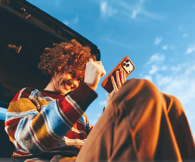 Smiling woman looking at her phone while sitting on the trunk of her vehicle.