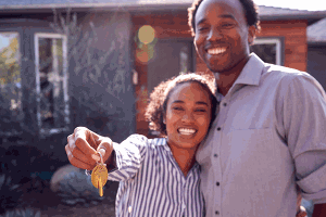 A smiling couple holding house keys in front of their first home.