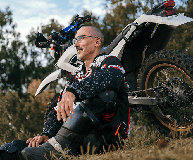 Man sitting next to his dirt bike and smiling outside.