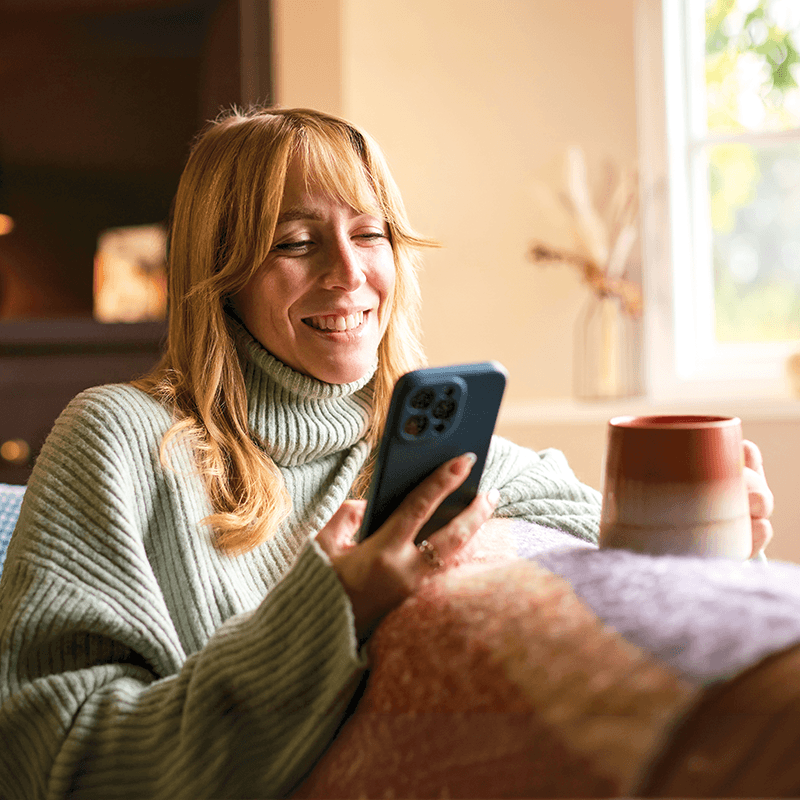 Happy woman sitting on the couch and smiling looking at her phone with a mug.