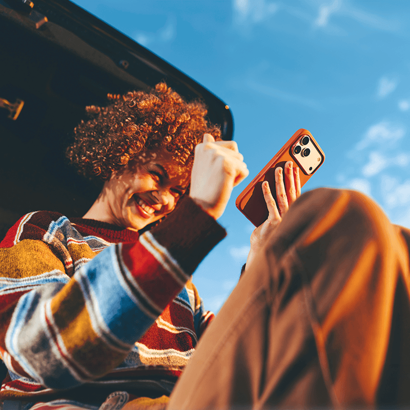 Happy woman smiling on her phone while sitting on her trunk of vehicle.
